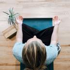 person sitting on blue yoga mat in cross-legged position on wooden floor with plant and yoga blocks next to them